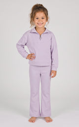 Toddler girl wearing a lavender half-zip sweatshirt and matching flared pants, standing barefoot.