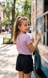 Girl wearing a lilac short-sleeve crew-neck tee with relaxed black shorts, standing beside a display board.