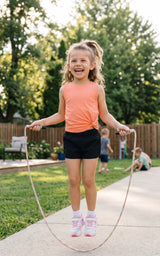Girl wearing a coral sleeveless tank with black shorts, jumping rope outdoors.