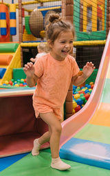 Toddler girl wearing a coral pink terry short-sleeve top and matching dolphin shorts in a play area.