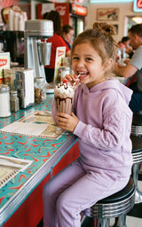 Toddler girl wearing a lavender brushed-terry hoodie and tapered joggers, holding a milkshake at a diner.