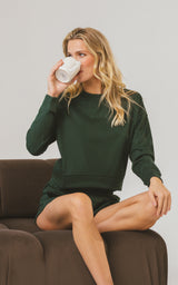 Woman wearing a dark pine green ribbed long-sleeve lounge top and matching shorts, seated on a brown sofa with a mug.