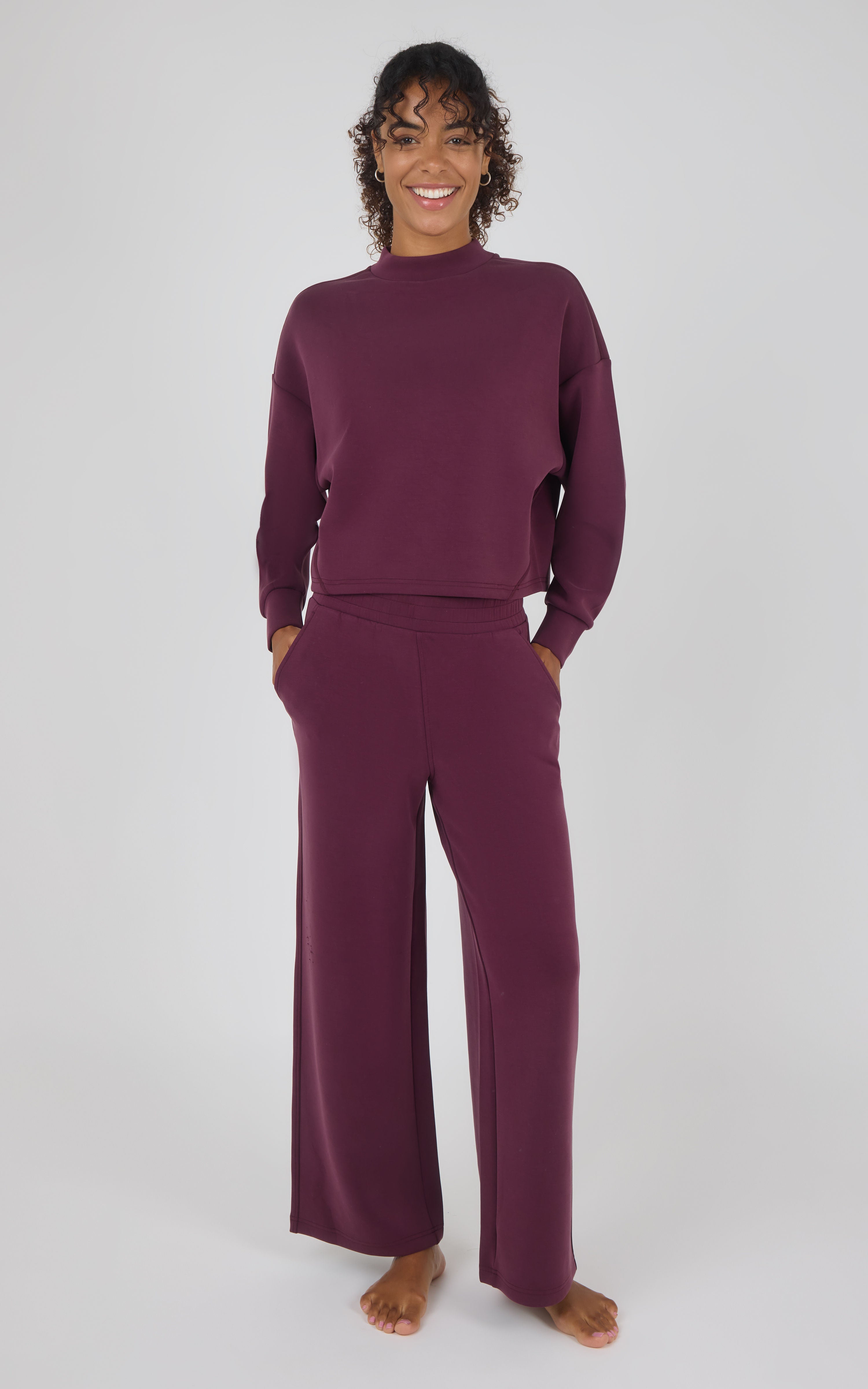 A woman modeling a matching two-piece loungewear set in deep plum purple, featuring a long-sleeve mock neck top and wide-leg pants, against a plain light gray background.