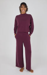 A woman modeling a matching two-piece loungewear set in deep plum purple, featuring a long-sleeve mock neck top and wide-leg pants, against a plain light gray background.