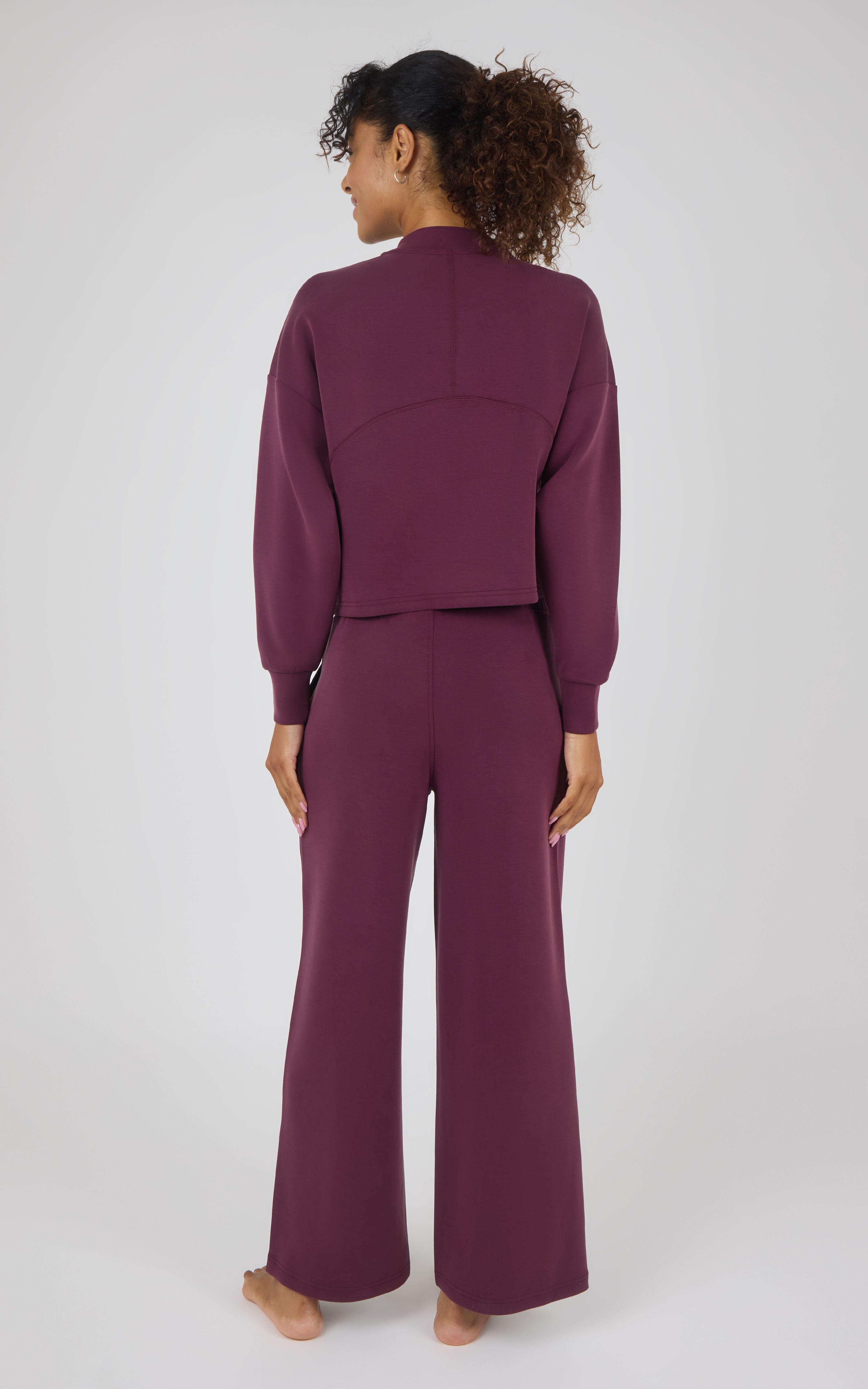 A woman stands barefoot against a light gray studio background, viewed from the back, wearing a matching mauve-purple cropped long-sleeve top and wide-leg pants set.