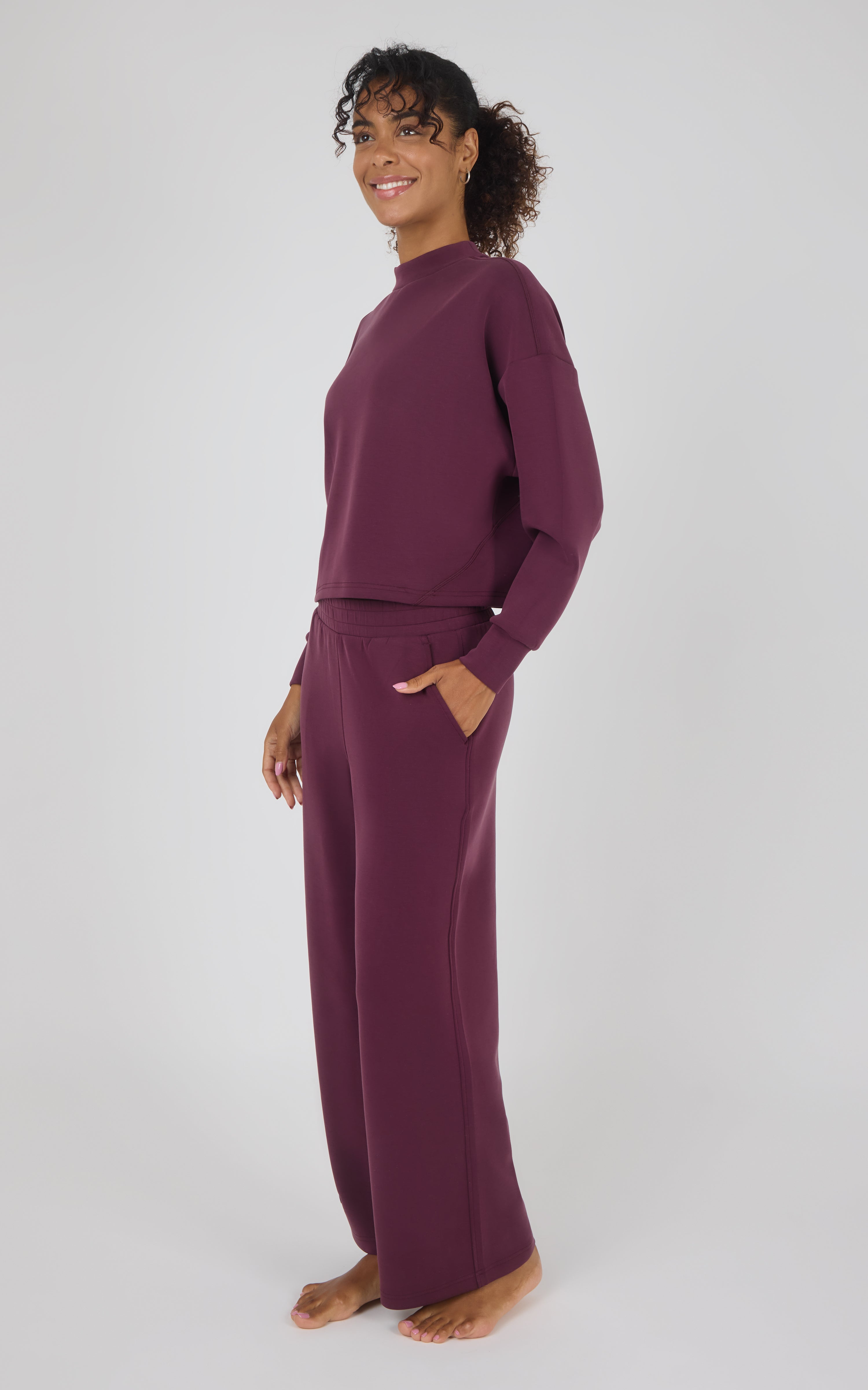 A woman modeling a matching two-piece loungewear set in a deep plum purple color, consisting of a long-sleeve mock neck top and wide-leg pants, against a plain light gray background.