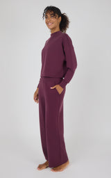 A woman modeling a matching two-piece loungewear set in a deep plum purple color, consisting of a long-sleeve mock neck top and wide-leg pants, against a plain light gray background.