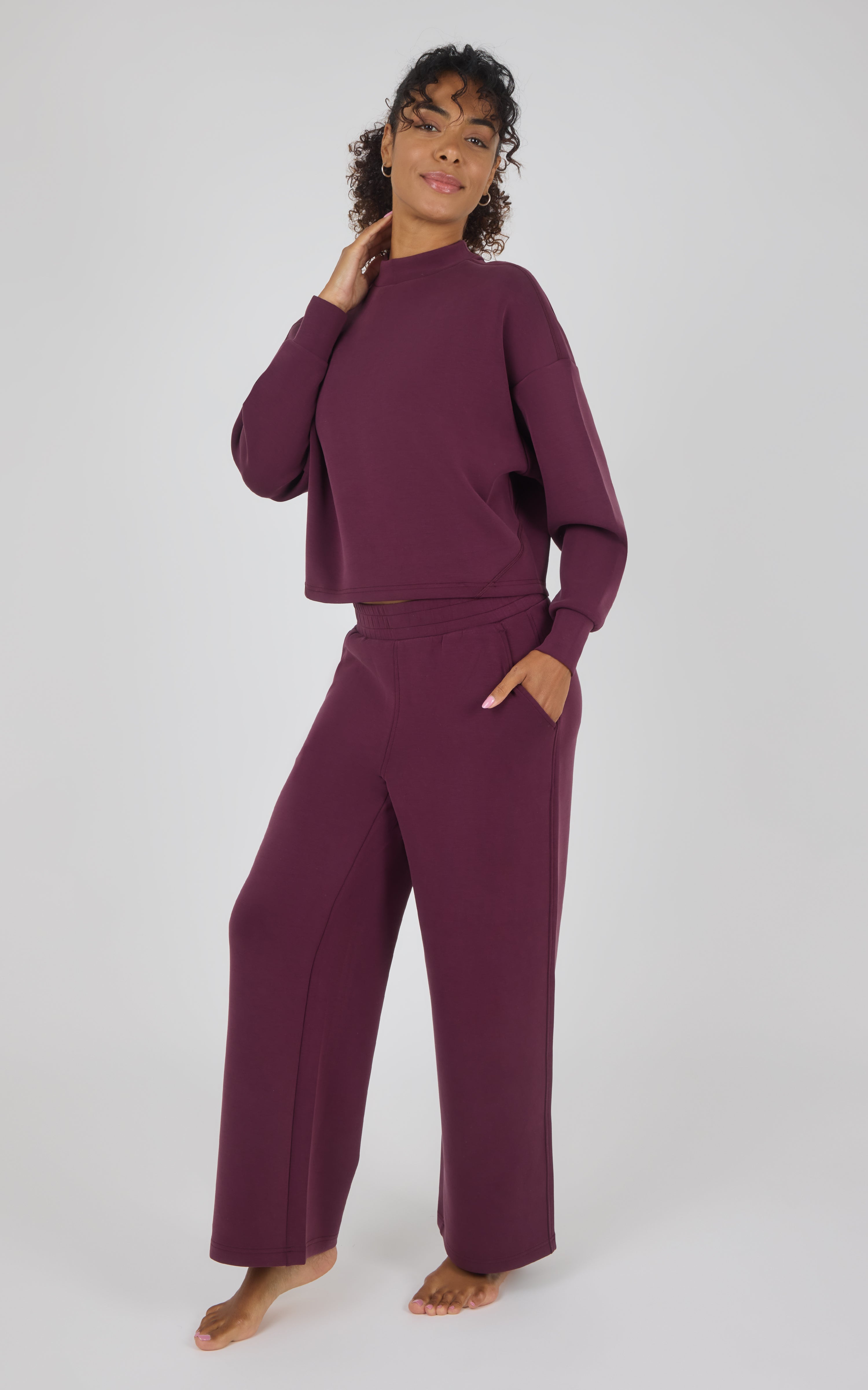 A woman modeling a matching two-piece loungewear set in deep plum purple, featuring a long-sleeve cropped top and wide-leg pants, against a light gray studio background.