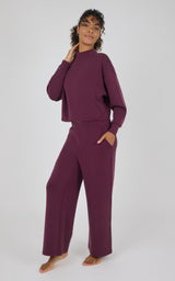 A woman modeling a matching two-piece loungewear set in deep plum purple, featuring a long-sleeve cropped top and wide-leg pants, against a light gray studio background.