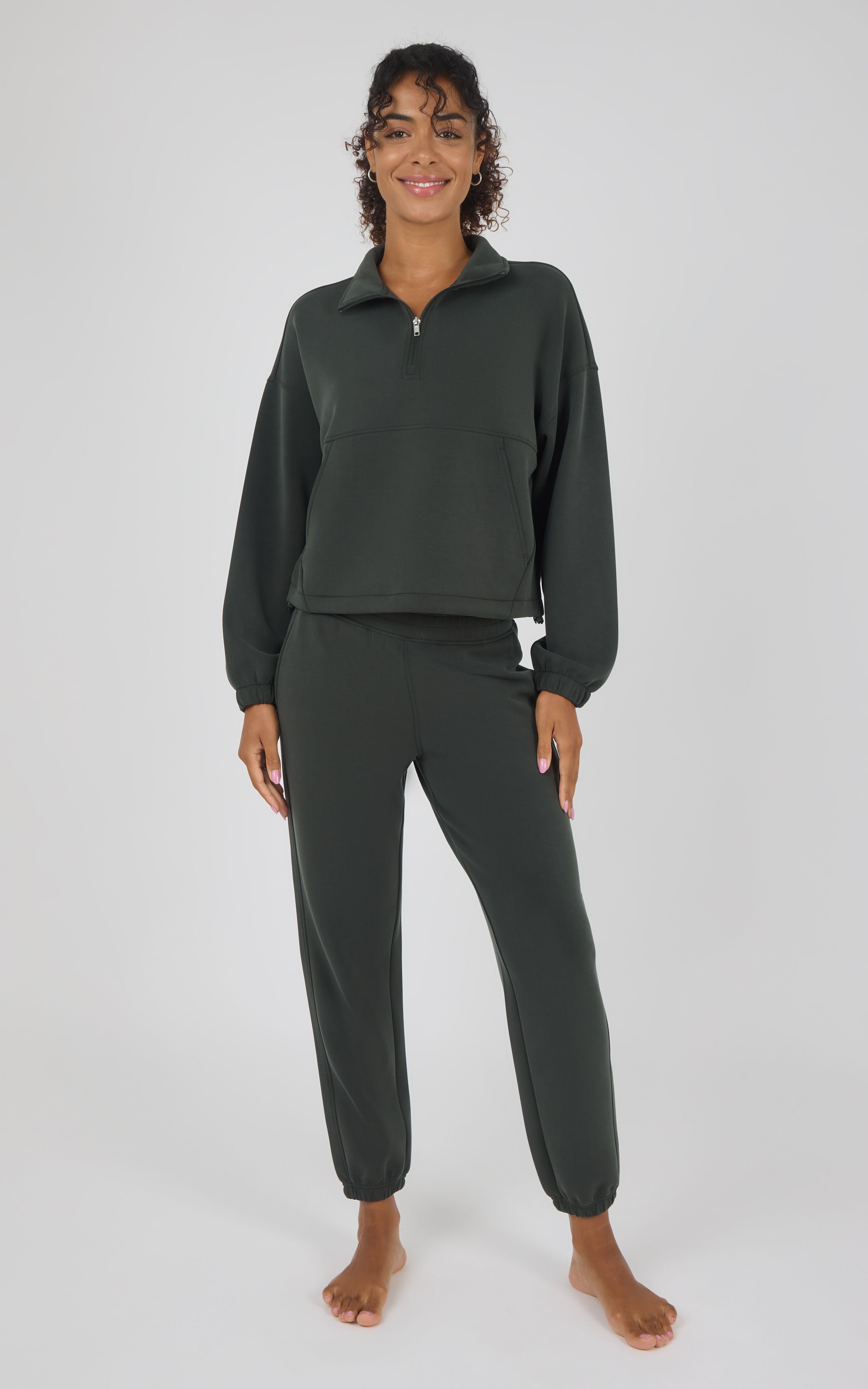 A woman modeling a dark olive green two-piece loungewear set consisting of a cropped quarter-zip long-sleeve top and matching jogger pants, shot against a light gray studio background.