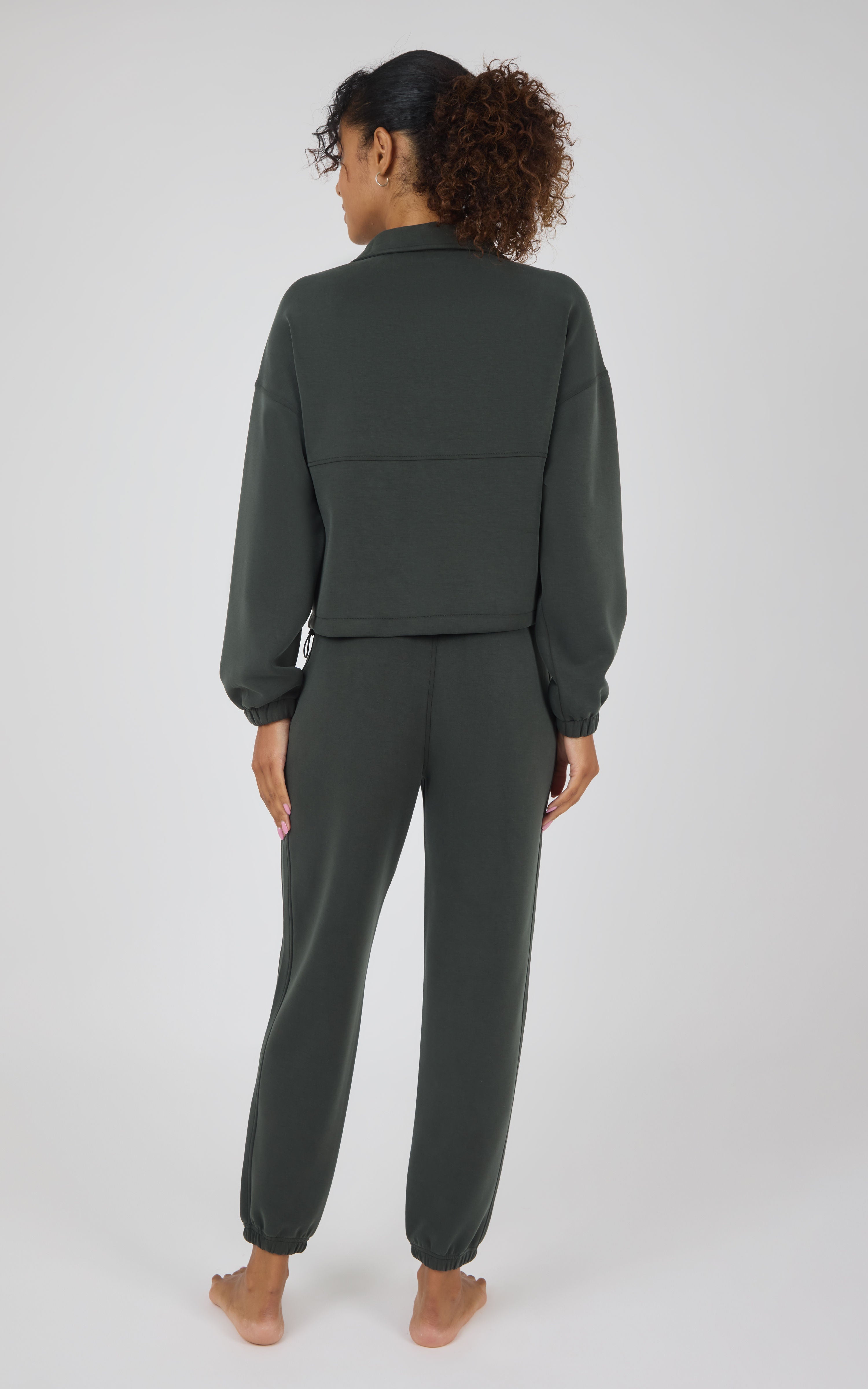 A woman viewed from the back wearing a dark olive green matching lounge set consisting of a cropped long-sleeve top and jogger pants, standing barefoot against a light gray studio background.