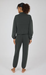 A woman viewed from the back wearing a dark olive green matching lounge set consisting of a cropped long-sleeve top and jogger pants, standing barefoot against a light gray studio background.