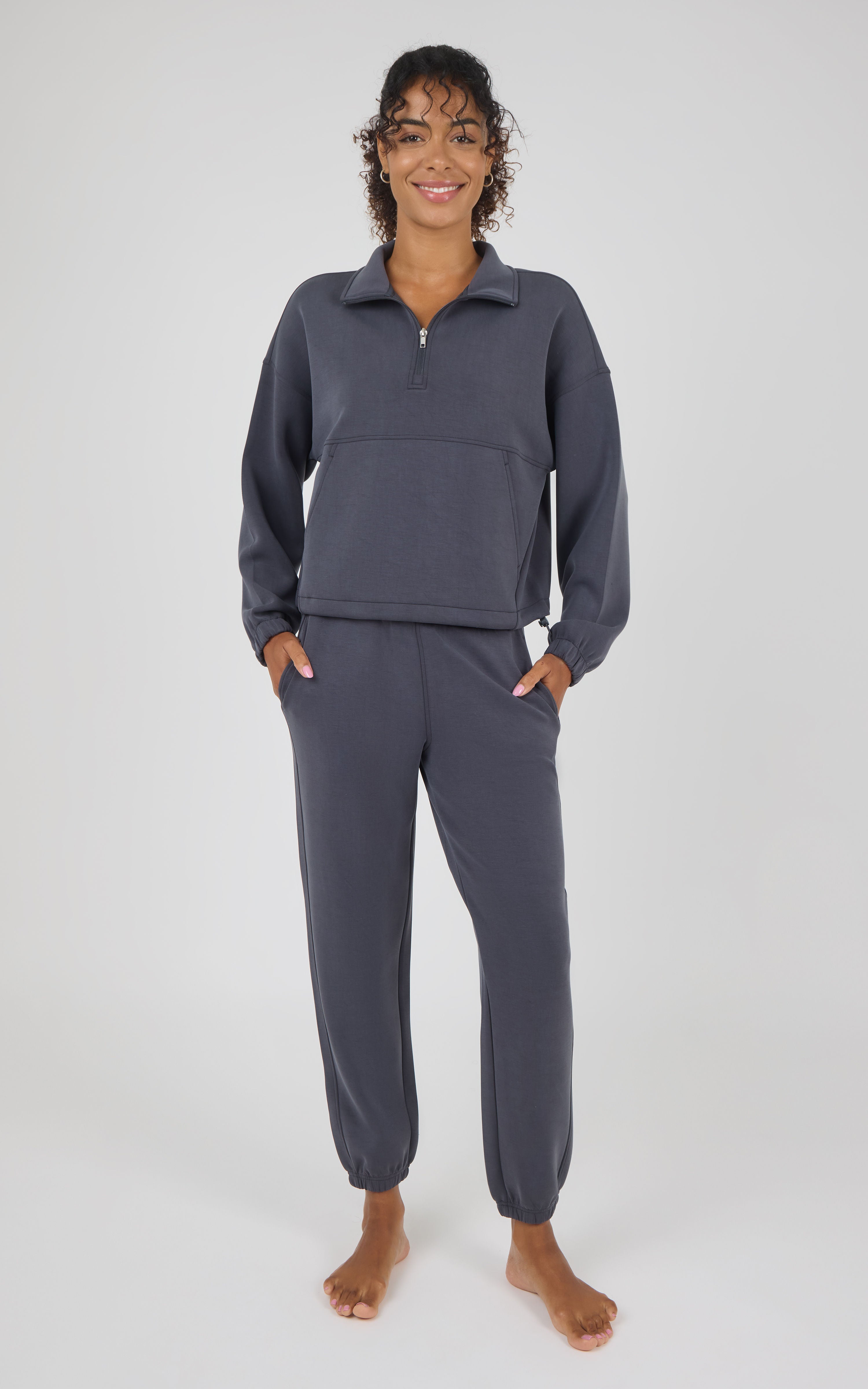 A woman modeling a matching dark gray two-piece loungewear set consisting of a quarter-zip long-sleeve top and matching jogger pants, standing against a light gray studio background.