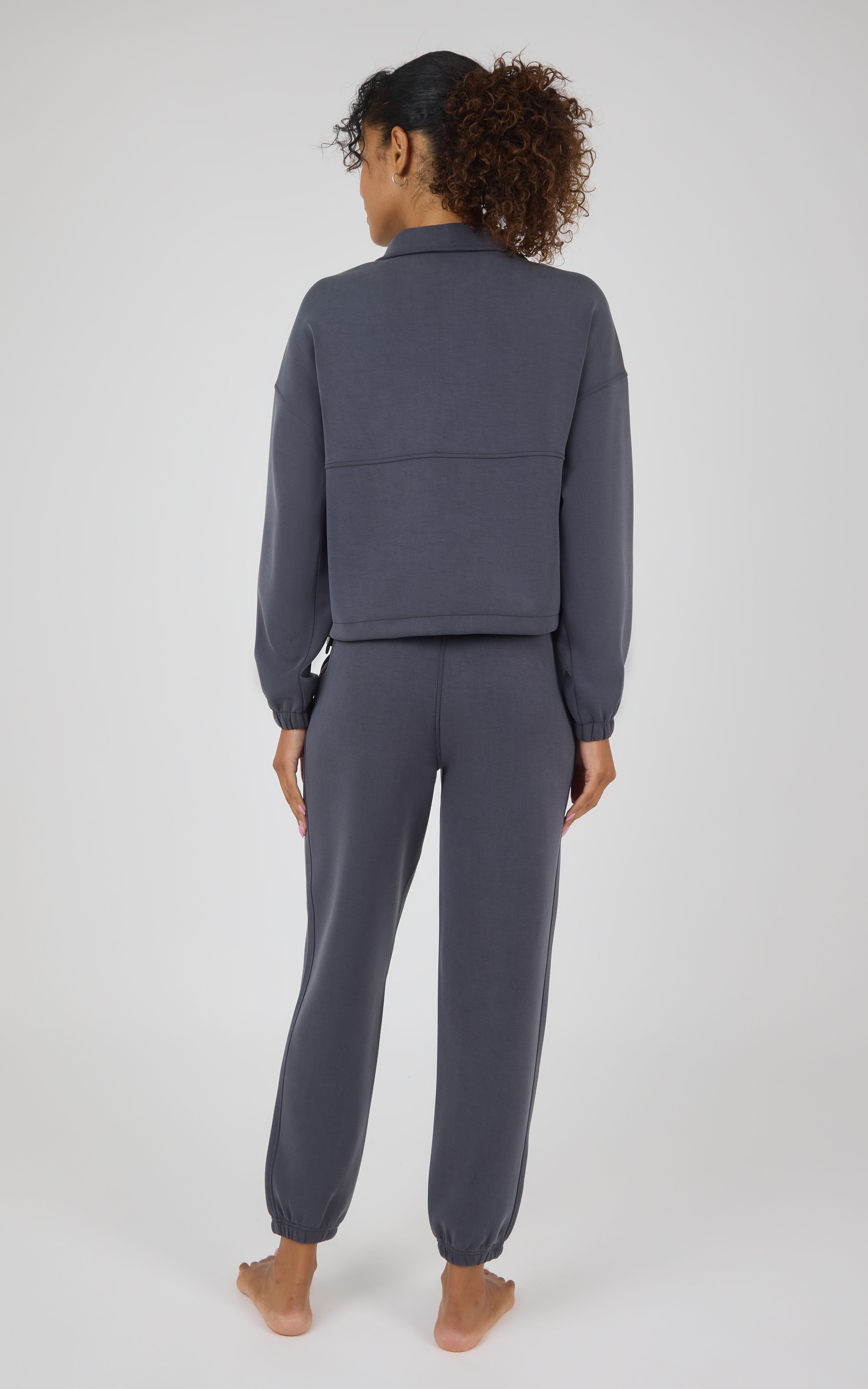 A woman viewed from behind wearing a matching dark slate gray lounge set consisting of a cropped long-sleeve top and jogger pants, standing barefoot against a light gray studio background.
