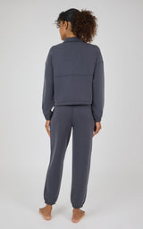 A woman viewed from behind wearing a matching dark slate gray lounge set consisting of a cropped long-sleeve top and jogger pants, standing barefoot against a light gray studio background.