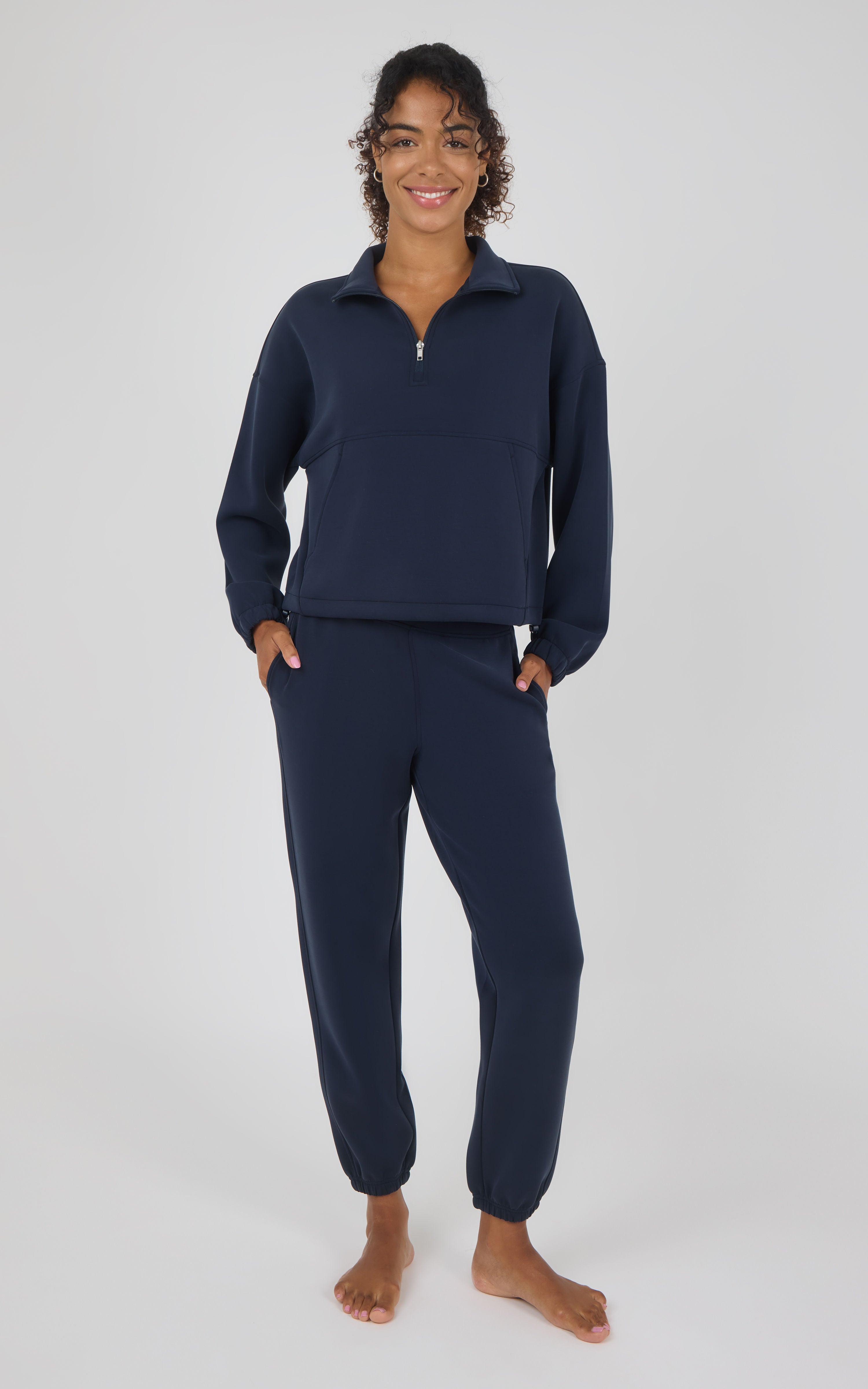 A woman modeling a matching navy blue two-piece loungewear set, consisting of a quarter-zip long-sleeve top and coordinating jogger pants, against a light gray studio background.