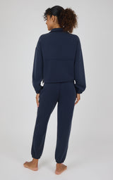 A woman is shown from behind wearing a matching navy blue lounge set consisting of a cropped long-sleeve top and jogger pants, standing barefoot against a light gray studio background.