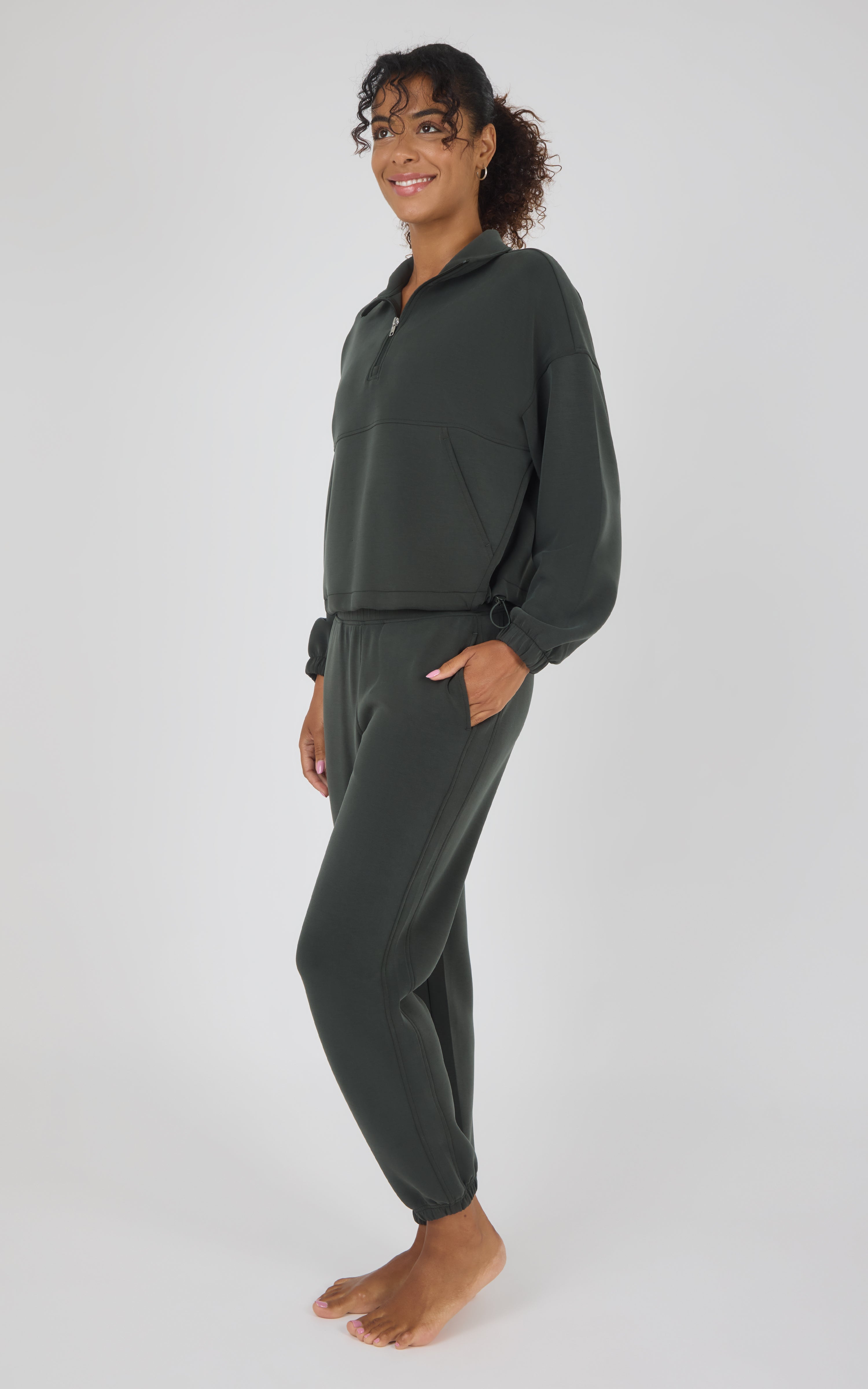 A woman modeling a dark olive green two-piece loungewear set consisting of a half-zip pullover and matching jogger pants, standing against a light gray studio background.