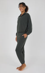 A woman modeling a dark olive green two-piece loungewear set consisting of a half-zip pullover and matching jogger pants, standing against a light gray studio background.