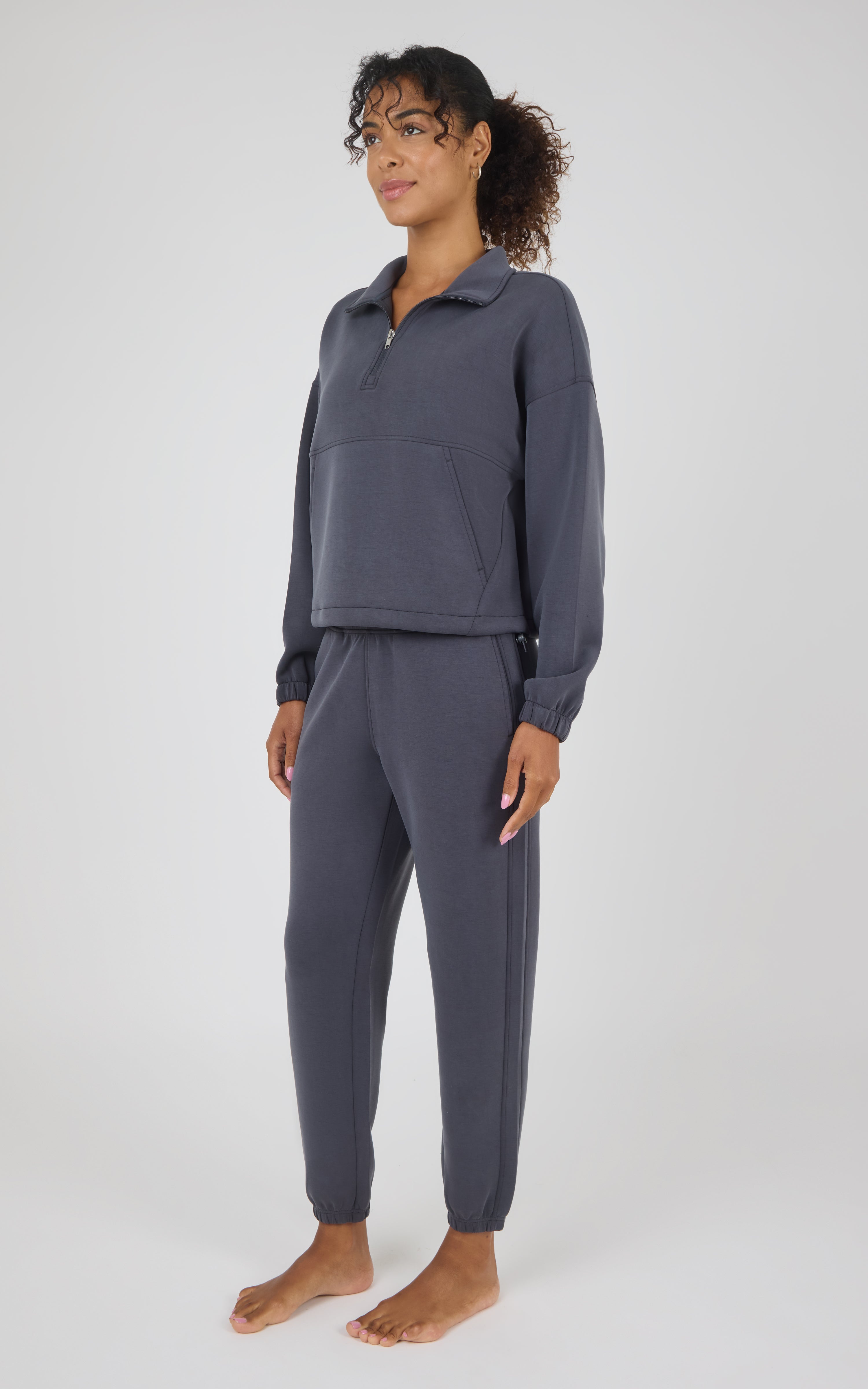 A woman modeling a matching dark slate gray lounge set consisting of a long-sleeve quarter-zip pullover and matching jogger pants, standing against a light gray studio background.