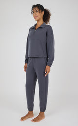 A woman modeling a matching dark slate gray lounge set consisting of a long-sleeve quarter-zip pullover and matching jogger pants, standing against a light gray studio background.