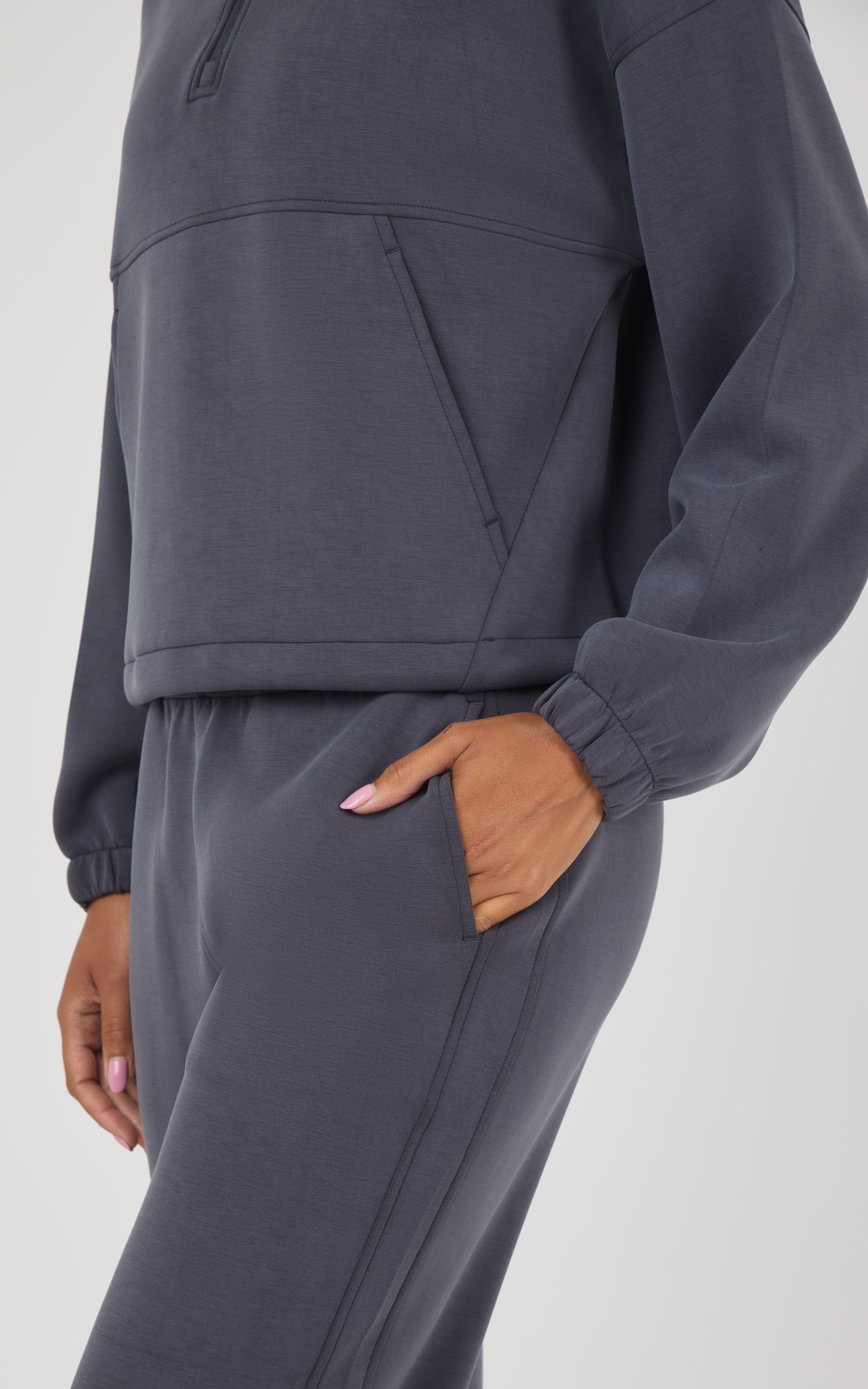 A close-up of a person wearing a dark gray matching lounge set, featuring a cropped quarter-zip sweatshirt and matching jogger pants with side pockets.