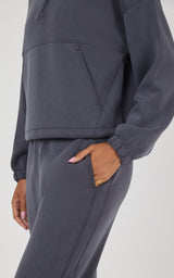 A close-up of a person wearing a dark gray matching lounge set, featuring a cropped quarter-zip sweatshirt and matching jogger pants with side pockets.
