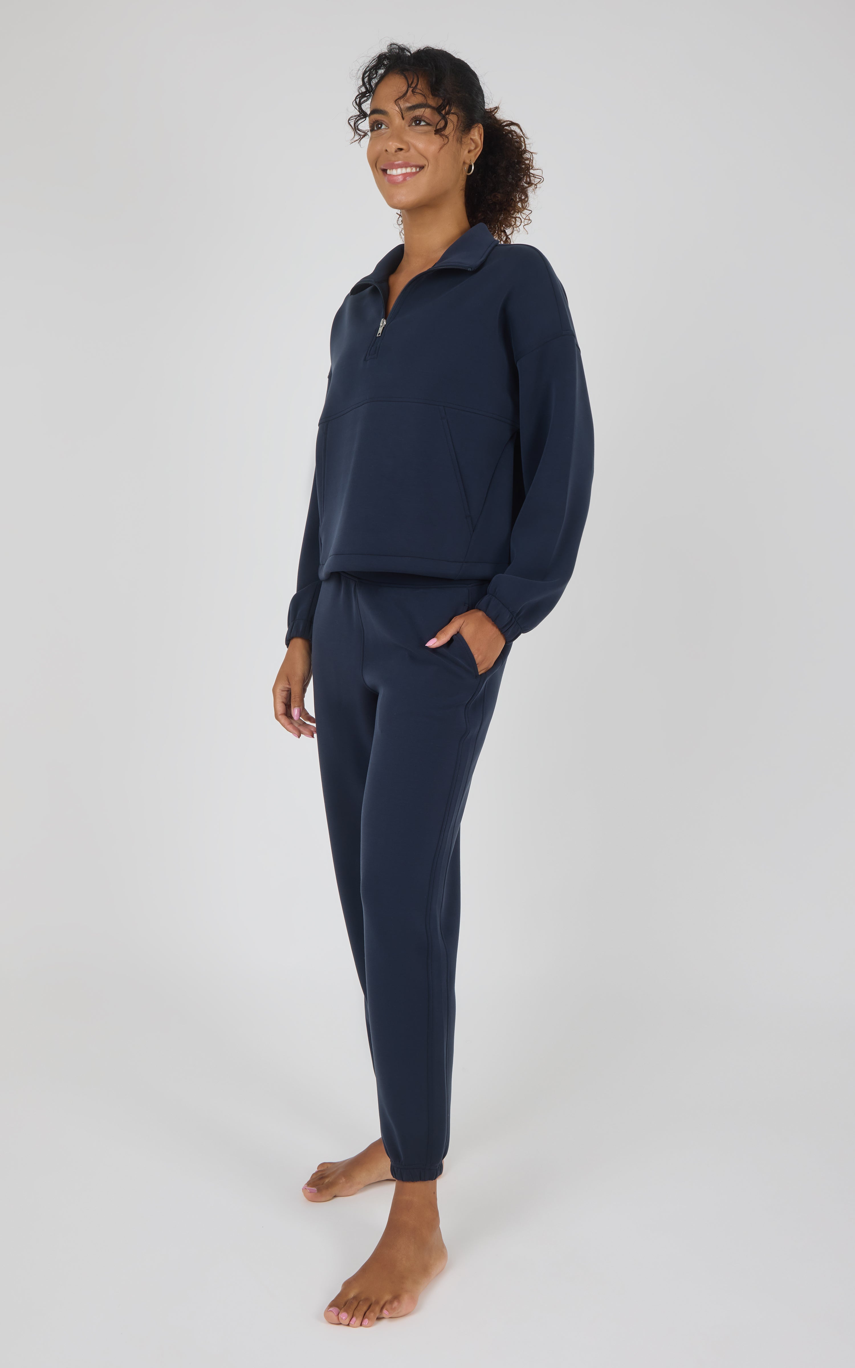 A woman modeling a matching navy blue loungewear set consisting of a quarter-zip long-sleeve top and coordinating pants, standing against a light gray studio background.