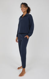 A woman modeling a matching navy blue loungewear set consisting of a quarter-zip long-sleeve top and coordinating pants, standing against a light gray studio background.