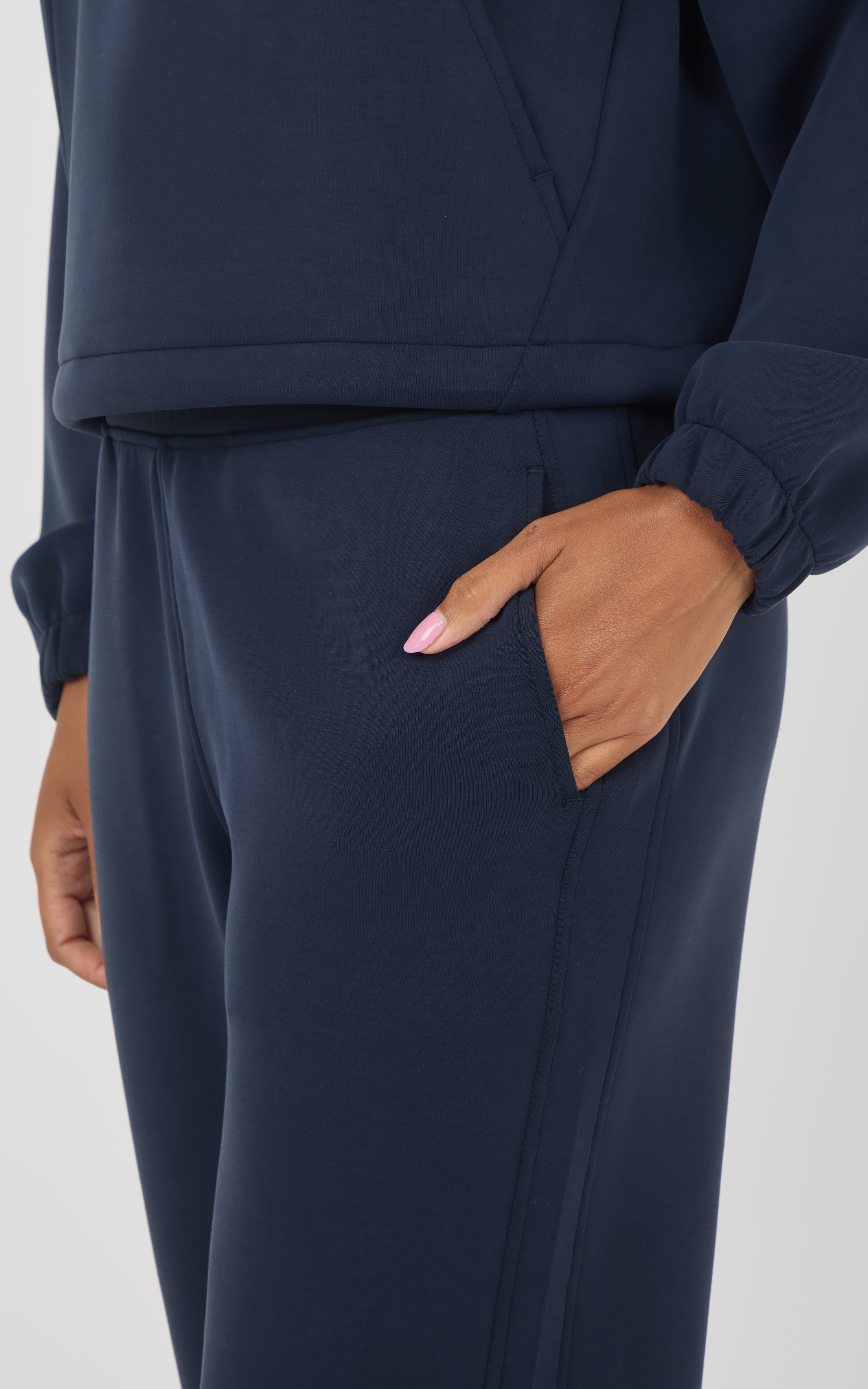 Close-up of a person wearing a matching navy blue tracksuit, showing the side pocket of the pants with one hand in the pocket, against a light gray studio background.
