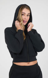 Woman wearing a black cropped zip-up hoodie with the hood up and matching black pants.