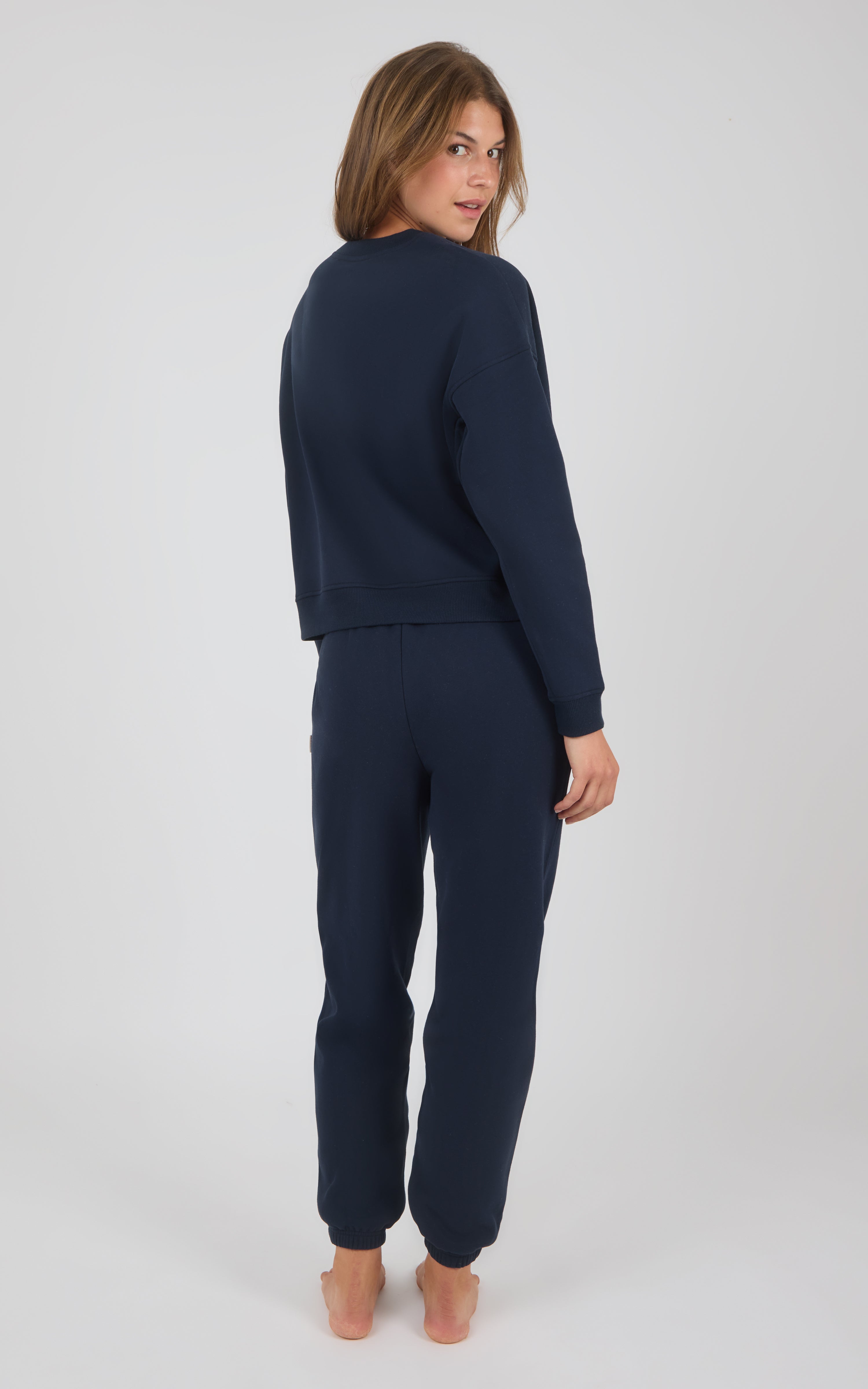 Woman wearing navy blue long-sleeve top and matching pintuck jogger pants, barefoot, standing against gray background.