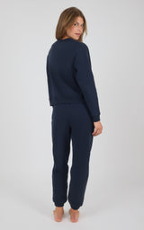 Woman wearing navy blue long-sleeve top and matching pintuck jogger pants, barefoot, standing against gray background.