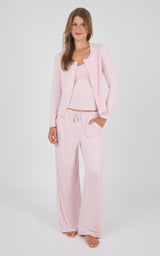 A woman modeling a light pink loungewear set consisting of a cropped tank top, matching wide-leg pants with a drawstring waist, and a long-sleeve button-front cardigan, against a plain light gray studio background.