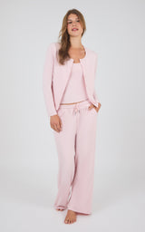 A woman modeling a light pink loungewear set consisting of a cropped cardigan, a scoop-neck tank top, and matching wide-leg drawstring pants, shot against a plain light gray studio background.