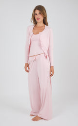 A woman modeling a light pink loungewear set consisting of a cropped tank top, a matching open-front cardigan, and wide-leg pants, shot against a plain light gray studio background.
