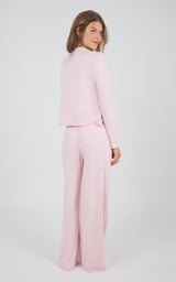 A woman modeling a light pink two-piece outfit consisting of a cropped long-sleeve top and matching wide-leg trousers, photographed against a plain light gray studio background.