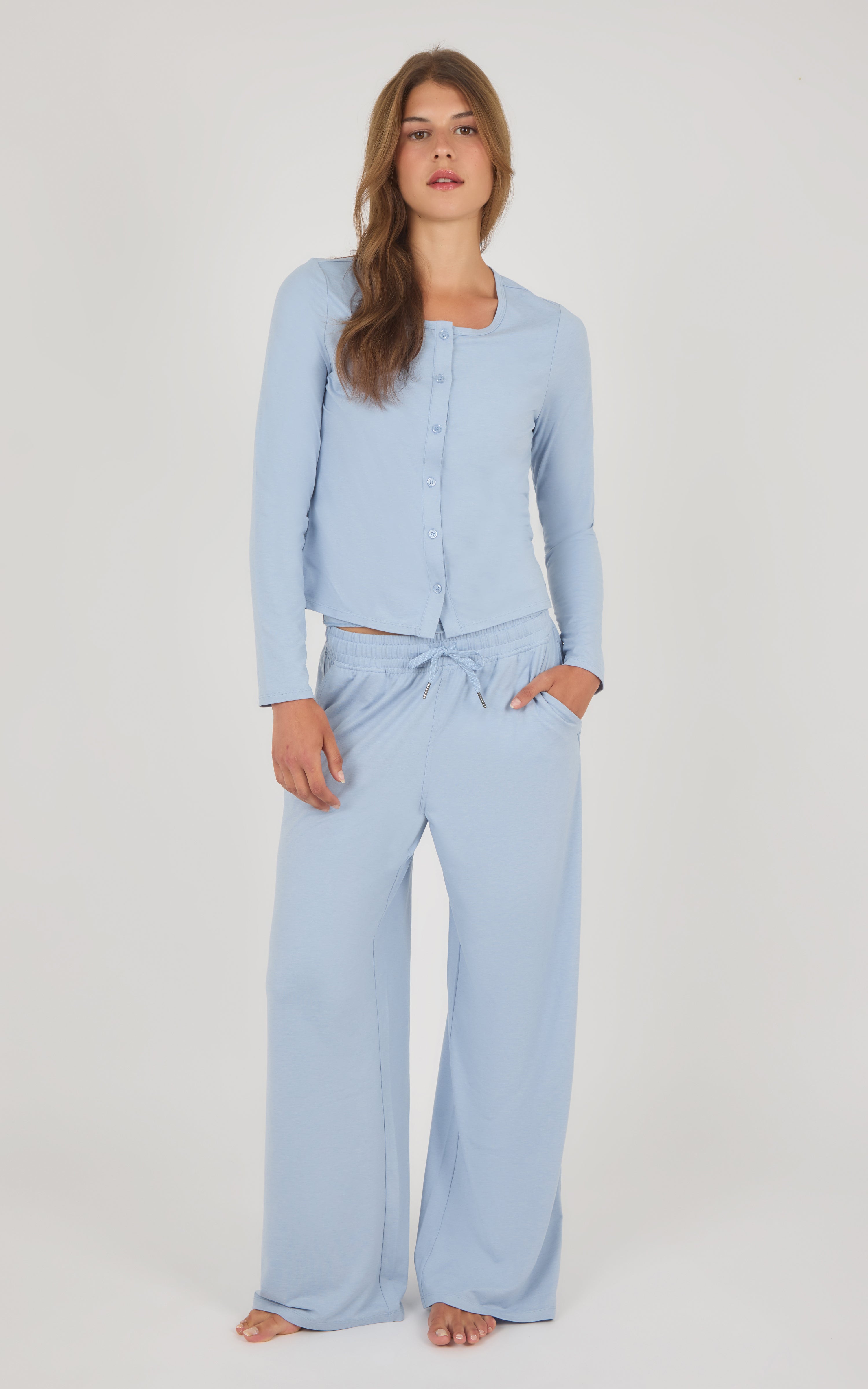 A woman modeling a light blue two-piece loungewear set consisting of a long-sleeve button-front top and matching wide-leg pants with a drawstring waist, against a plain light gray studio background.