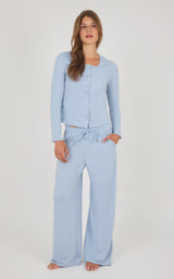 A woman modeling a light blue two-piece loungewear set consisting of a long-sleeve button-front top and matching wide-leg pants with a drawstring waist, against a plain light gray studio background.