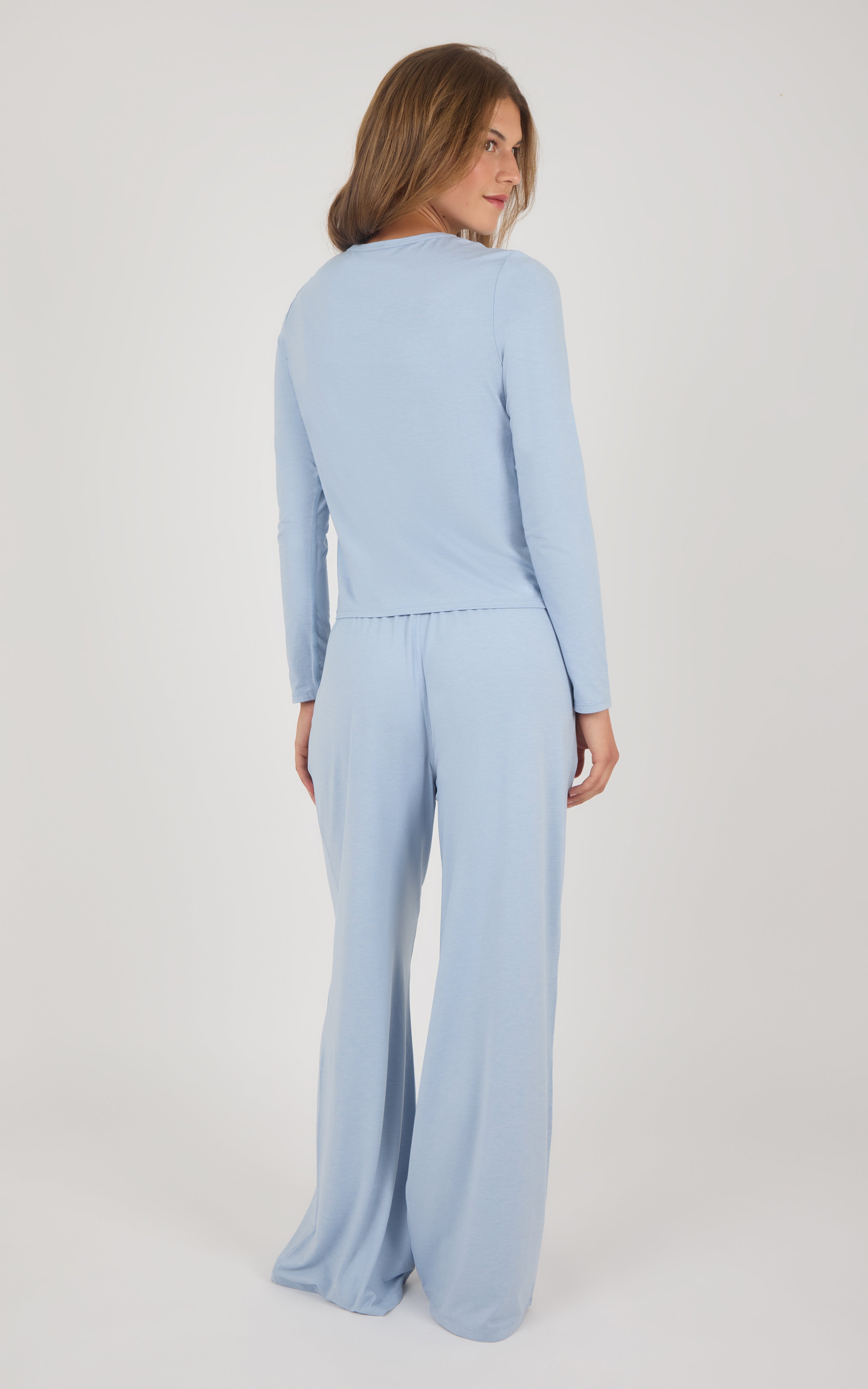 A woman wearing a light blue long-sleeve jumpsuit, photographed from the back against a plain light gray studio background.