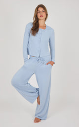 A woman modeling a light blue two-piece loungewear set consisting of a long-sleeve button-front top and matching wide-leg pants, photographed against a plain light gray studio background.