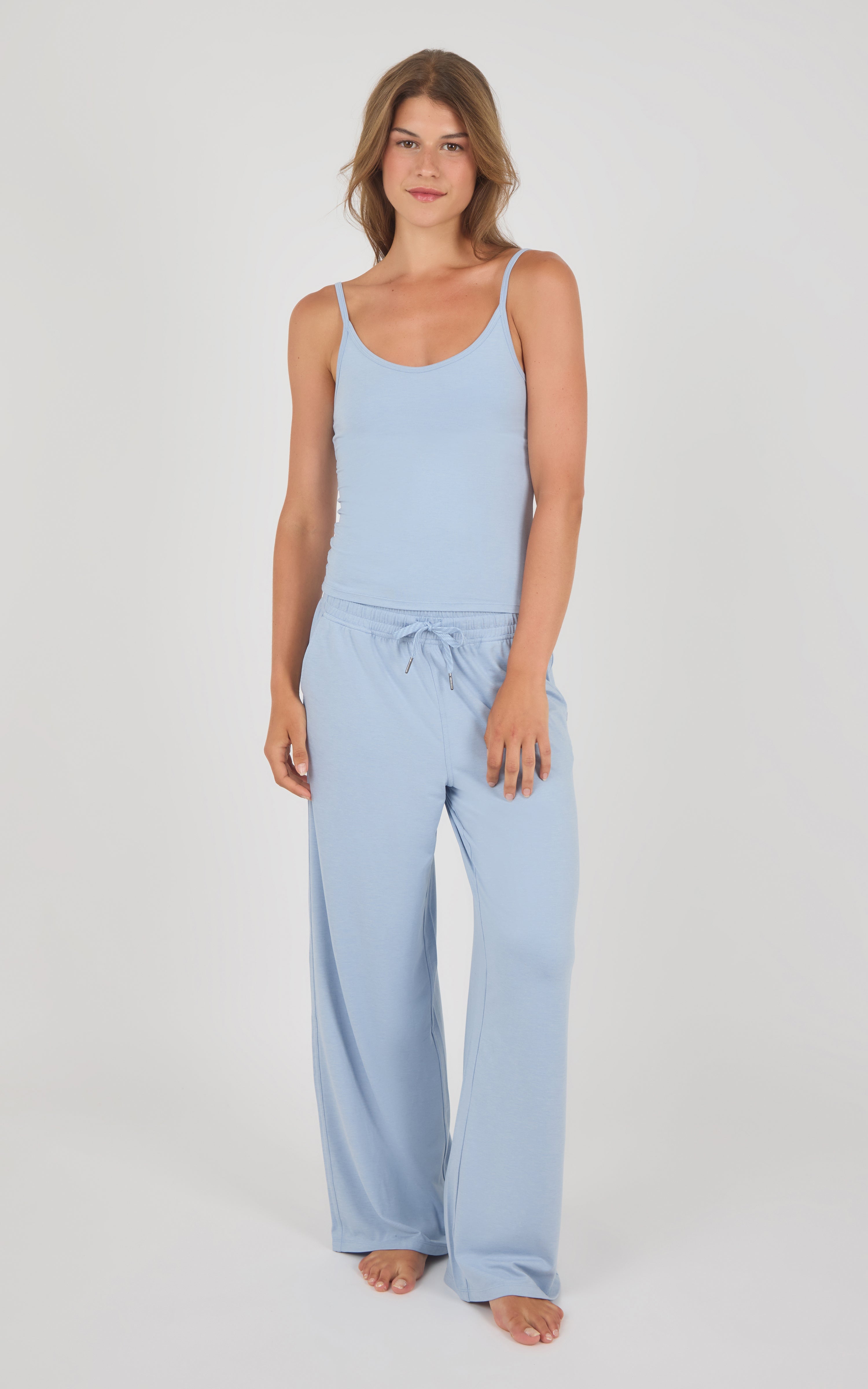 A woman modeling a light blue two-piece loungewear set consisting of a sleeveless camisole top and matching wide-leg drawstring pants, shot against a neutral light gray studio background.