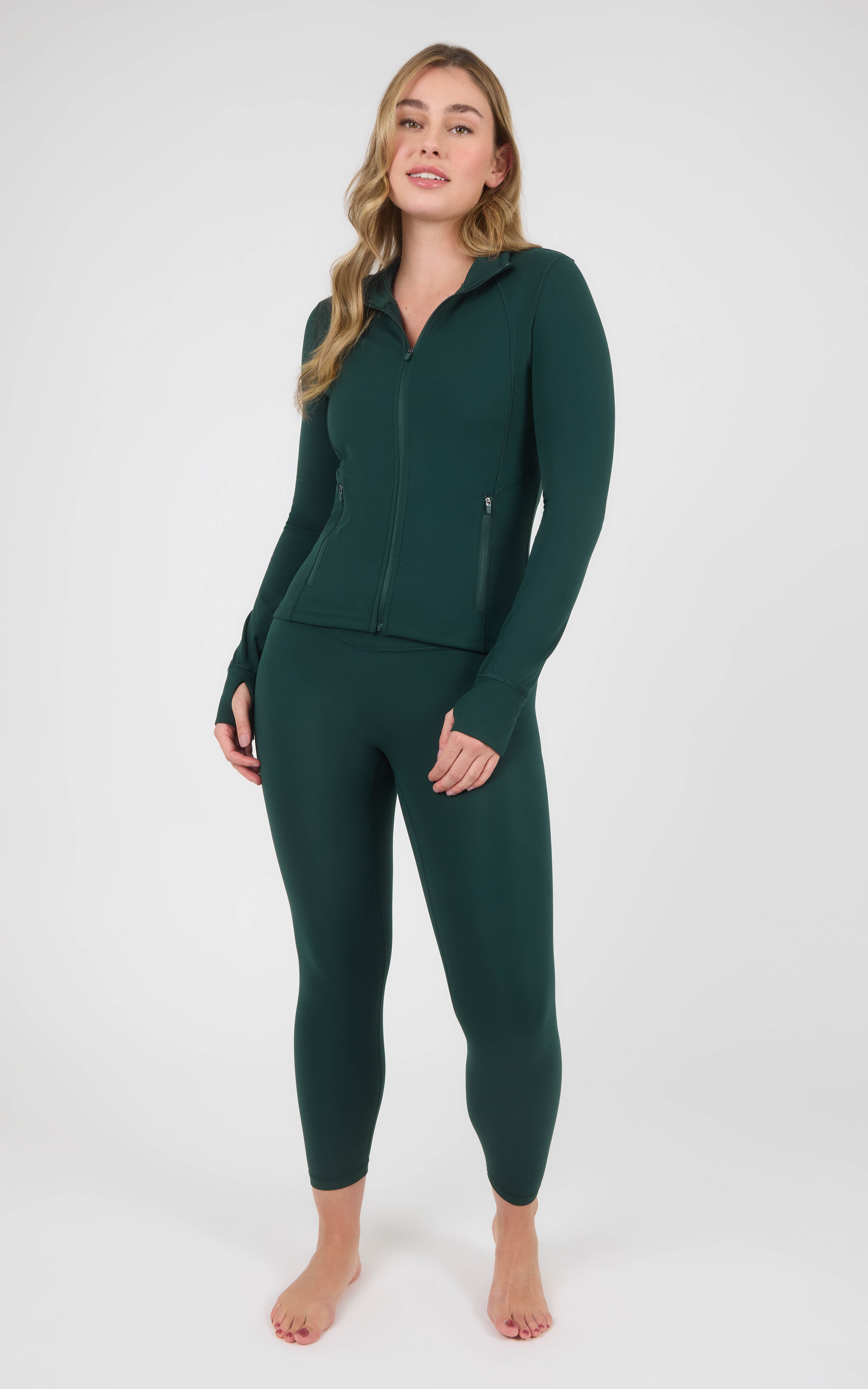 Woman wearing a dark forest-green zip-up jacket and matching high-waisted ankle leggings, shown full-length against a plain.