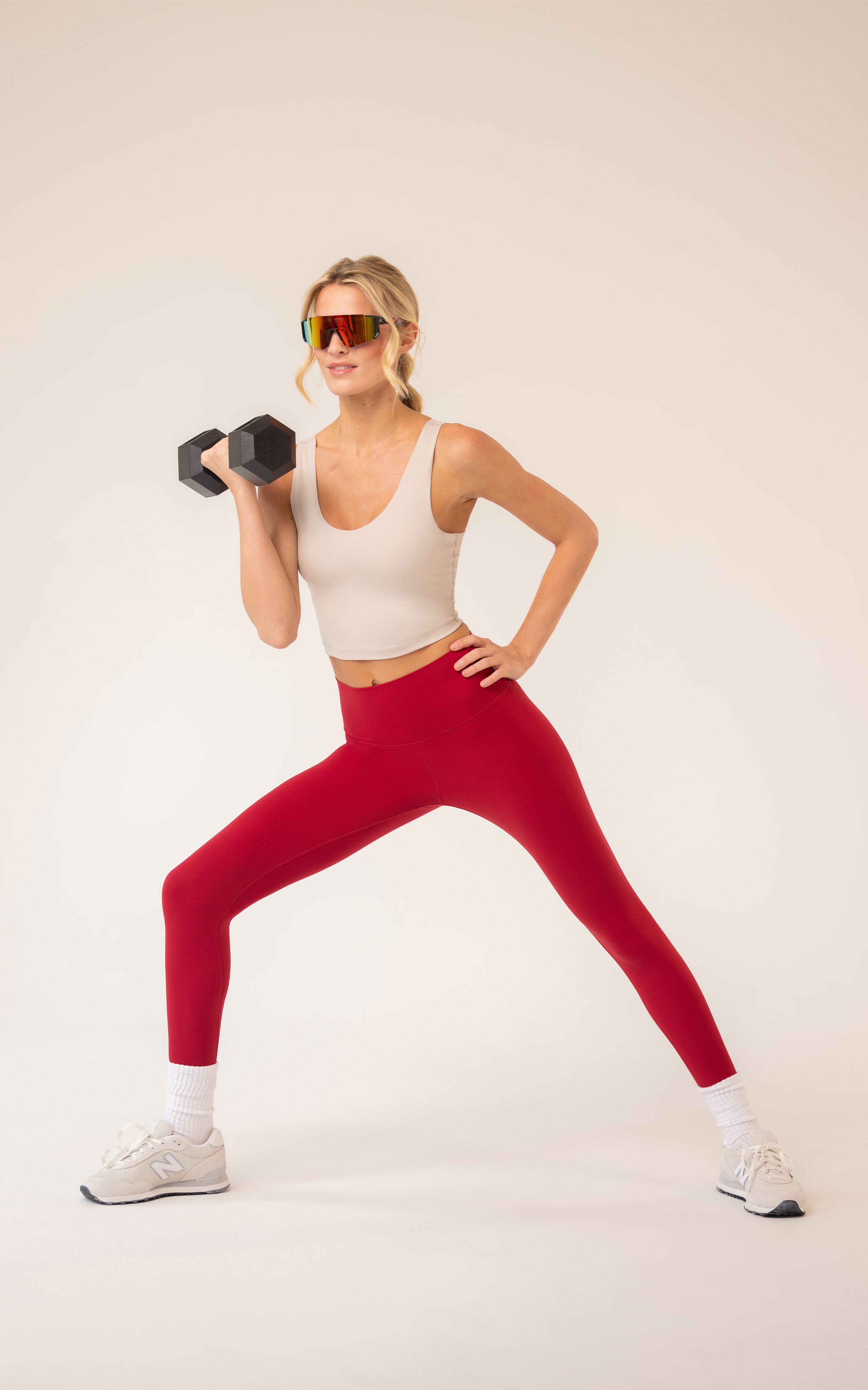 Woman wearing a white scoop-neck tank top with a built-in bra and bright red leggings, holding a dumbbell in a wide lunge.