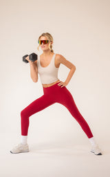 Woman wearing a white scoop-neck tank top with a built-in bra and bright red leggings, holding a dumbbell in a wide lunge.