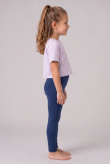 Little girl in profile wearing full-length navy leggings and a lavender short-sleeve top, standing barefoot.