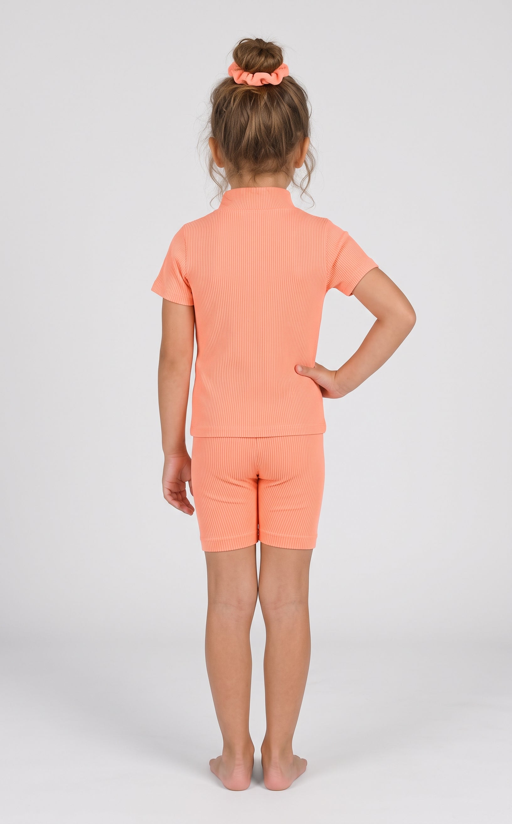 Toddler girl modeling a coral ribbed short-sleeve top and matching fitted shorts, shown from behind.