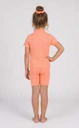 Toddler girl modeling a coral ribbed short-sleeve top and matching fitted shorts, shown from behind.