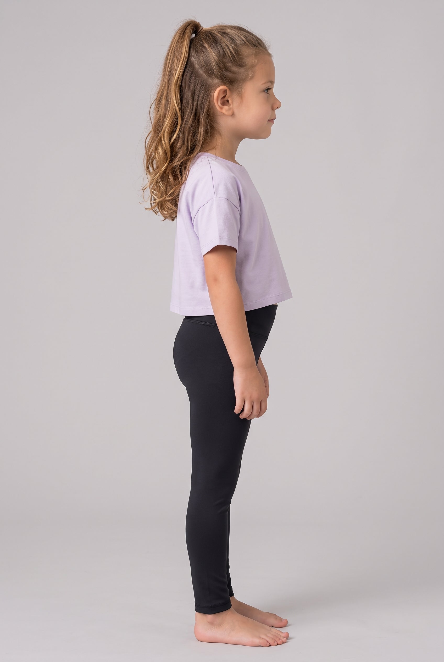 Little girl in profile wearing full-length black leggings and a lavender T-shirt, standing barefoot.