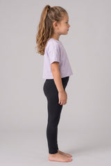 Little girl in profile wearing full-length black leggings and a lavender T-shirt, standing barefoot.